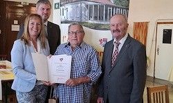 Ein Mann hält ein Zertifikat in der Hand, während er zwischen zwei Frauen und einem weiteren Mann steht, alle posieren drinnen in der Nähe eines Tisches.