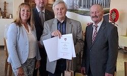Vier Menschen stehen eng beieinander in einem Raum, wobei zwei Männer ein Zertifikat halten. Sie alle lächeln und eine Frau schaut den Mann an, der das Zertifikat hält.