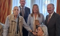 Eine Gruppe von Menschen steht in formeller Kleidung zusammen, ein älterer Mann hält ein Buch, eine Frau eine Trophäe, und andere lächeln in einem Raum.