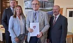 Vier Menschen in formeller Kleidung stehen zusammen, einer der Männer hält ein Zertifikat. Sie posieren für ein Foto. Im Hintergrund befinden sich ein Tisch und Stühle.