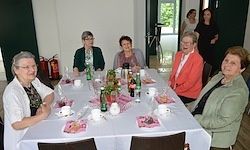 Eine Gruppe von Frauen sitzt an einem Tisch mit Essen und Getränken. Sie lächeln und posieren für ein Foto. Der Tisch ist mit Tassen, Tellern und Flaschen gedeckt. Dahinter befindet sich eine Tür zu einem anderen Raum.
