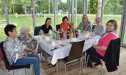 Eine Gruppe älterer Frauen sitzt um einen Tisch in einem Essbereich, mit einem grünen Feld im Hintergrund.