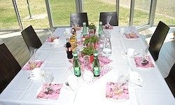 Ein Esstisch ist mit Flaschen, Tassen, Tellern und Besteck gedeckt. Er ist mit einer weißen Tischdecke bedeckt und mit rosa Tischsets verziert.