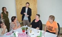 Ein Mann steht und führt einen Zaubertrick vor einer Gruppe von Frauen vor, die um einen Tisch mit Weinflaschen, Weingläsern und einem Topf sitzen.