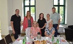 Eine Gruppe von Menschen posiert für ein Foto an einem Tisch mit Essen, Getränken und Weingläsern. Sie stehen vor einer Wand mit Fenstern und Bäumen draußen.