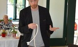 Ein Mann in einem schwarzen Blazer und grauem Hemd demonstriert einen Knoten in einem Raum. Hinter ihm sitzt eine Frau an einem Tisch mit Pflanzen und Kerzen.