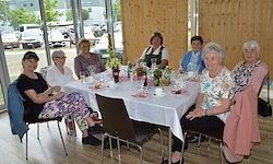 Eine Gruppe älterer Frauen sitzt um einen Tisch, möglicherweise zu einem Familientreffen. Sie tragen formelle Kleidung und einige tragen Brillen. Der Tisch ist mit Weingläsern, Flaschen und Tellern gedeckt.