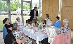 Eine Gruppe älterer Frauen sitzt um einen Esstisch mit Weinflaschen, Tellern und Tassen. Ein Mann steht hinter ihnen, möglicherweise im Gespräch. Der Raum hat Holzwände, eine Glastür und Blick auf geparkte Autos.