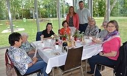 Eine Gruppe von Menschen, einschließlich älterer Frauen, versammelt sich um einen Tisch, möglicherweise für ein Treffen, mit Blick auf eine grüne Wiese draußen.