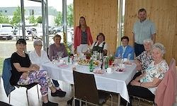 Eine Gruppe älterer Frauen und Männer versammelt sich um einen Tisch für eine Mahlzeit, mit einer Person im Hintergrund.