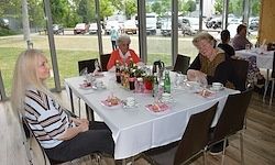 Vier Frauen sitzen an einem Tisch in einem Restaurant. Der Tisch ist für eine Mahlzeit mit Tellern, Tassen und verschiedenen Flaschen gedeckt. Das Restaurant hat Glaswände, und draußen gibt es einen Parkplatz mit mehreren Autos und einem Rasenbereich.