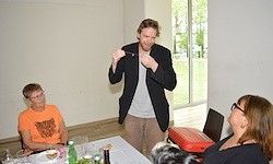 Ein Mann in einem schwarzen Blazer steht und gestikuliert vor einer sitzenden Gruppe am Tisch, mit einem roten Koffer und Laptop in der Nähe, in einem Raum mit Glasfenstern.