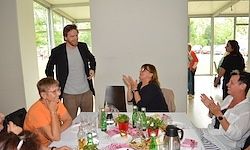 Eine Gruppe von Menschen sitzt um einen Tisch mit Flaschen und Essen. Ein Mann im Anzug steht hinter ihnen und schaut einen anderen Mann an. Zwei Frauen klatschen. Glasfenster mit Blick auf einen Parkplatz und Bäume.