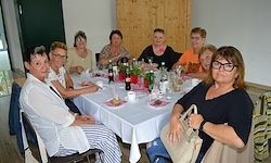 Eine Gruppe von Frauen sitzt um einen Tisch herum und genießt eine Mahlzeit mit verschiedenen Flaschen und Tellern. Die Umgebung wirkt ungezwungen, mit einer Holzwand und einem Fenster im Hintergrund.