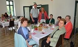 Eine Gruppe älterer Frauen und Männer versammelt sich um einen Tisch in einem Speisesaal, lächelt und posiert für ein Foto. Sie sind von Tischen und Stühlen umgeben, mit ein paar Leuten, die hinter ihnen stehen. Der Raum hat Holzböden und weiße Wände.