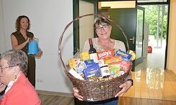 Eine Frau hält einen Korb voller Snacks und Getränke, lächelt und steht in einem Raum. Eine andere Frau hält einen blauen Becher hinter ihr, und eine ältere Frau schaut zu. Eine Tür steht offen, mit Bäumen draußen.