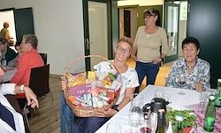 Eine Frau hält einen Korb mit Essen an einem Tisch, an dem andere Menschen sitzen und stehen. Eine Person hält ein Glas Wein.