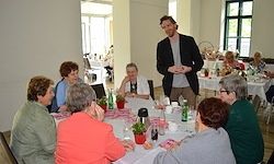 Ein Mann steht vor einer Gruppe sitzender Frauen in einem Raum mit weißen Wänden, Tischen und Pflanzen. Sie scheinen ein Gespräch zu führen.