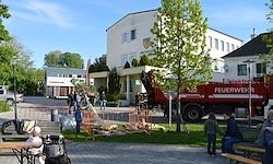 Ein Außenbereich mit mehreren versammelten Personen, einem roten Feuerwehrwagen auf der Straße und einem weißen Gebäude im Hintergrund.