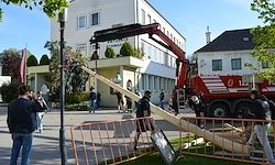 Menschen versammeln sich an einem Baugelände, wo ein großer Holzbalken von einem Kran vor einem Gebäude angehoben wird.