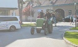 Ein grüner Traktor fährt mit Menschen darauf die Straße hinunter. Ein weißer Van ist geparkt in der Nähe, und mehrere Menschen laufen auf dem Bürgersteig. Dahinter steht ein Gebäude mit einem Baum und einer Straßenlaterne.