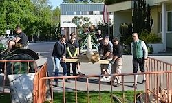Eine Gruppe von Menschen bewegt einen großen Holzstamm vor einem Gebäude, während andere zuschauen und eine Person ein Fahrrad fährt.