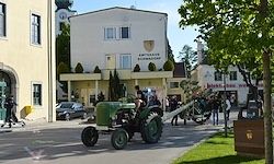 Ein grüner Traktor fährt durch einen Stadtplatz mit einem Gebäude im Hintergrund. Menschen gehen und stehen in der Umgebung.
