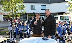 Zwei Männer stehen an einem Tisch mit einem Mikrofon. Sie sind von einer Gruppe von Menschen in blauen Uniformen umgeben. Die Veranstaltung scheint im Freien stattzufinden, mit Gebäuden und Bäumen im Hintergrund.