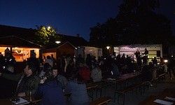 Eine Menschenmenge versammelt sich nachts in einem Essbereich im Freien, Tische sind mit Lichtern aufgestellt und eine Bühne ist in der Ferne aufgebaut.