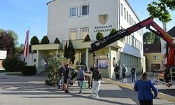 Ein Gebäude mit mehreren Fenstern und Fahnen, mit einem Kran, der etwas anhebt. Mehrere Menschen stehen davor, einige tragen Kisten.