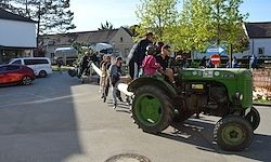 Eine Gruppe von Kindern und Erwachsenen fährt auf einem grünen Traktor auf einem Parkplatz. Im Hintergrund sind Gebäude, Autos und Bäume zu sehen.