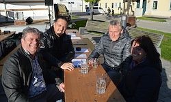Vier Personen sitzen um einen Holztisch mit Gläsern und Papieren. Sie befinden sich in einem Außenbereich mit Gebäuden und Bänken im Hintergrund.