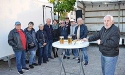 Eine Gruppe von Menschen steht um einen Tisch mit Bier und Essen herum, in der Nähe eines weißen LKWs.