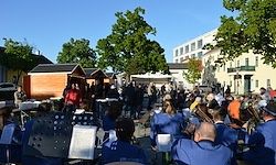Eine Band in blauen Uniformen spielt auf einem Stadtplatz, umringt von einer versammelten Menge. Bäume und Gebäude sind im Hintergrund zu sehen.