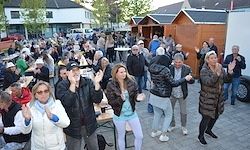 Eine große Gruppe von Menschen steht und tanzt in einem Außenbereich mit Tischen, Gebäuden und einem Fahrzeug im Hintergrund.