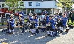 Eine Brassband spielt auf dem Pflaster vor einem Gebäude, mit einem zuschauenden Publikum. Die Bandmitglieder sitzen und stehen, halten verschiedene Blechblasinstrumente. Es gibt Bäume und einen Feuerwehrwagen im Hintergrund.