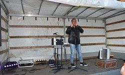 Ein Mann steht auf einer Bühne und singt in ein Mikrofon. Verschiedene Geräte sind auf dem Boden und auf einer kleinen Bühne platziert. Der Raum hat Holzwände und eine weiße Decke.