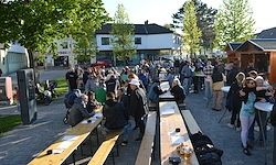Eine große Menschenmenge versammelt sich bei einer Veranstaltung im Freien, mit Menschen, die an langen Bänken unter Bäumen sitzen und Getränke und Essen genießen. Im Hintergrund sind Gebäude und Grünflächen zu sehen.