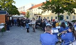 Eine Gruppe von Menschen in blauen Uniformen spielt Musikinstrumente in einem Freibereich. Viele Menschen stehen und sitzen vor ihnen. Bäume und Gebäude sind im Hintergrund.