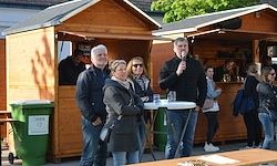 Ein Mann spricht in ein Mikrofon, während mehrere Menschen um Holzhütten herumstehen, möglicherweise bei einer Outdoor-Veranstaltung. Die Hütten befinden sich in der Nähe von grünen Mülleimern.