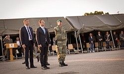 Drei Männer in Anzügen und einer in Uniform stehen vor einer Menschenmenge unter Zelten.