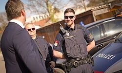 Drei Männer in Uniformen, einer mit Sonnenbrille, stehen neben Polizeifahrzeugen. Der Mann vorne trägt einen blauen Anzug und spricht mit einem Mann in schwarzer Uniform.