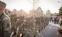 Eine Gruppe von Soldaten in Tarnuniformen marschiert mit Gewehre in der Hand die Straße entlang, während Zuschauer von den Seitenlinien zusehen.