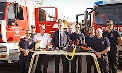Eine Gruppe Feuerwehrleute in Uniform, darunter zwei mit Krawatten, posieren für ein Foto vor Feuerwehrfahrzeugen. Sie halten Helme und Ausrüstung auf einem Tisch.