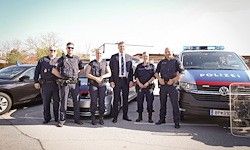 Eine Gruppe von Polizisten steht vor einem Polizeiwagen und zwei Autos, von denen eines ein Nummernschild hat. Sie tragen Uniformen und Sonnenbrillen.