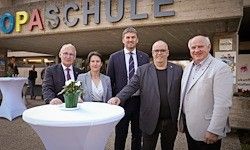 Fünf Personen stehen vor einem Gebäude, das mit 'OPASCHULE' beschriftet ist. Sie sind in Anzügen gekleidet. Ein weißer Tisch mit einer Blumenvase steht zwischen ihnen.