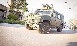 Ein gepanzertes Militärfahrzeug mit einem montierten Maschinengewehr fährt die Straße entlang. Eine Person geht im Hintergrund in der Nähe eines Gebäudes.