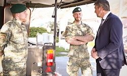 Drei Männer in Militäruniformen und Anzügen unterhalten sich im Freien. Zwei Soldaten stehen mit verschränkten Armen, während der Mann im Anzug aufmerksam zuhört.