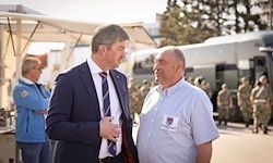 Zwei Männer in Geschäftskleidung unterhalten sich vor einem Bus. Hinter ihnen stehen Menschen und eine Frau in einer blauen Jacke. In der Ferne sind Soldaten in Uniform aneinandergereiht.