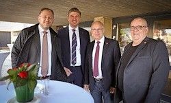 Vier Männer in formeller Kleidung stehen nebeneinander vor einem Tisch mit Blumenarrangement und posieren wahrscheinlich für ein Foto.